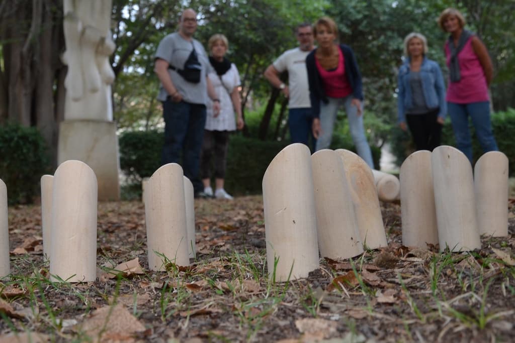 Douze quilles de Mölkky en bois posées en groupe sur un sol couvert de feuilles, avec un groupe de joueurs qui attend derrière.